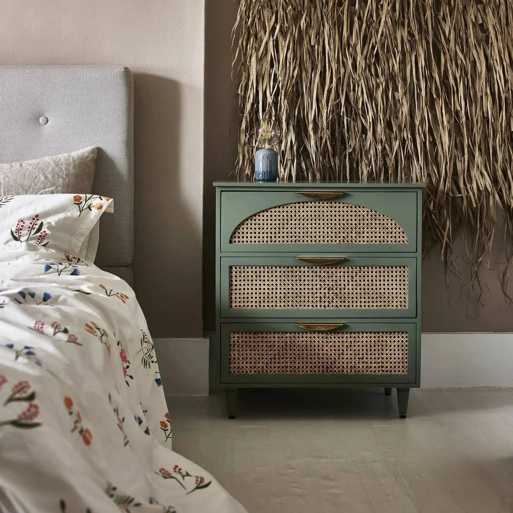 A green chest of drawers with rattan front panels and brass handles, placed in a bedroom setting.