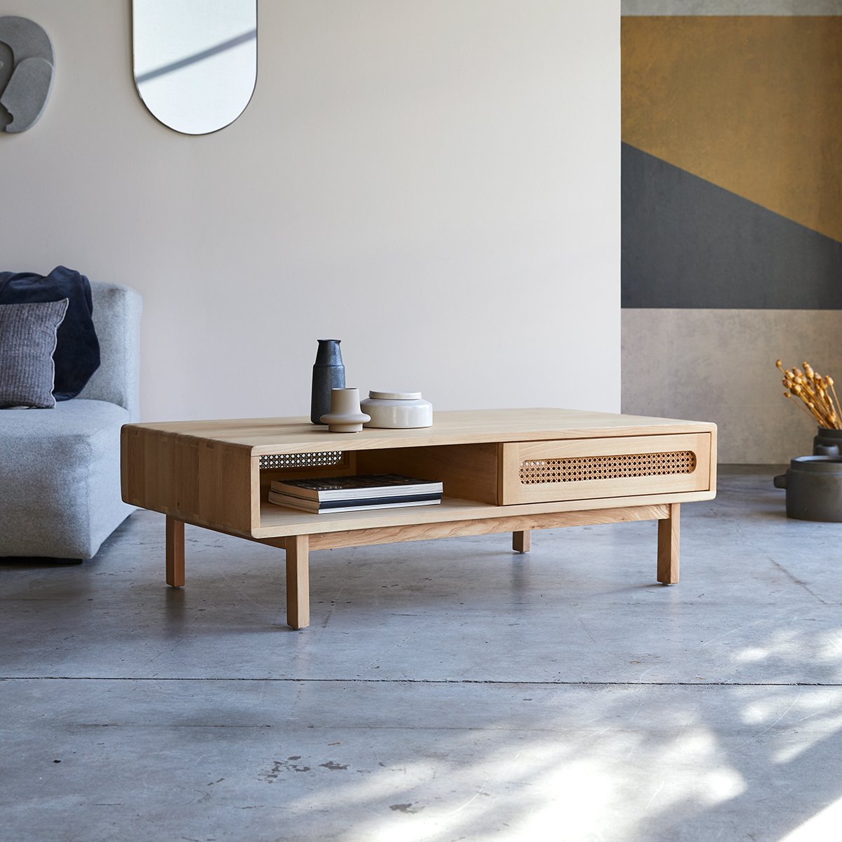 A rectangular Louis Coffee Table with natural rattan cane work, placed in a living room setting, with a plant, mirror, and furniture in the background.